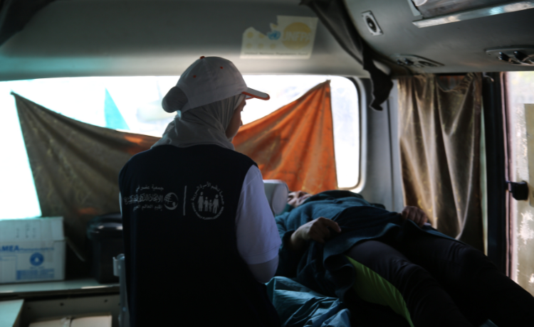 A female health worker in a blue vest and a white hat sits over a woman laying down inside a mobile clinic.